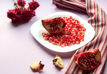  Cut pomegranate fruit with grains on a white plate, kitchen napkin, chrysanthemums on a purple background-the concept of eating a healthy vegetable