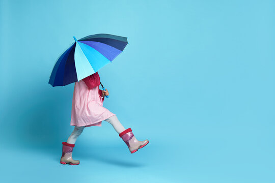 happy little child girl with blue umbrella in pink rain coat and rubber boots on blue background. copy space for text