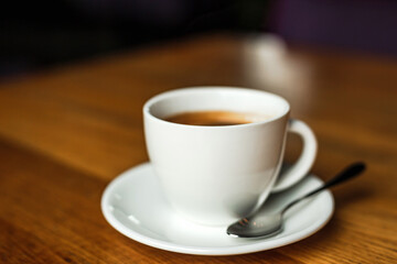 A white cup of black coffee, white saucer, spoon, wooden table, at a cafe