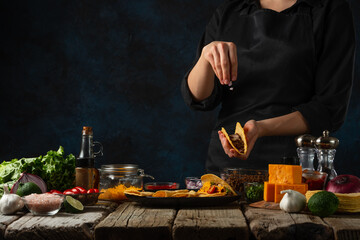 Professional chef in black uniform pours chopped garlic on corn tortilla for cooking tacos on dark blue background. Traditional mexican cuisine. Concept of tasty street food. Frozen motion.