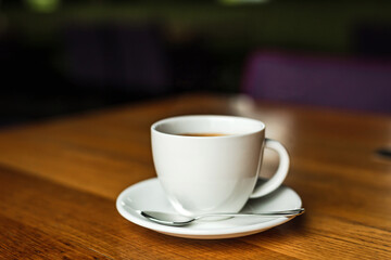 A white cup of black coffee, white saucer, spoon, wooden table, at a cafe