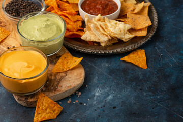 Top view of round metal plate with corn tortilla chips on dark blue background. Salsa, cheese dip and avocado souse for taste. Unhealthy food. Concept of spicy meal.