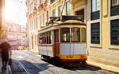 Naklejka na ściany i meble Lisbon Portugal. Yellow vintage tram driving by street of paving stones in district Alfama. Cityscape panorama with old houses and tower in sunny day with blue sky and white clouds.