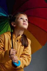 Cheerful child girl with multicolored umbrella in yellow rain coat on gray background.