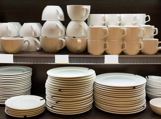 A stack of white plates are on the table. White coffee and tea cups are stacked on sale in the store.