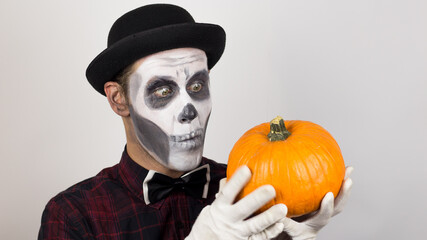 A horrible man in clown make-up holds a pumpkin, symbol of Halloween. A scary clown looks at the camera, holds a pumpkin in his hands and threatens her with a knife. Halloween costume.