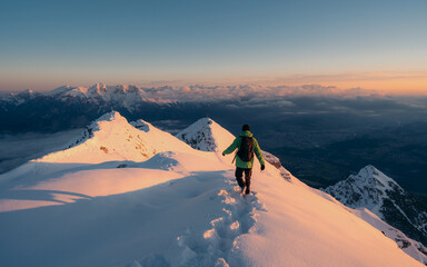 Top view of mountain peaks covered with snow, illuminated by sun hanging in distance with low cloud. A man in green jacket in his hand looking into the distance. Winter climbing, hiking 
