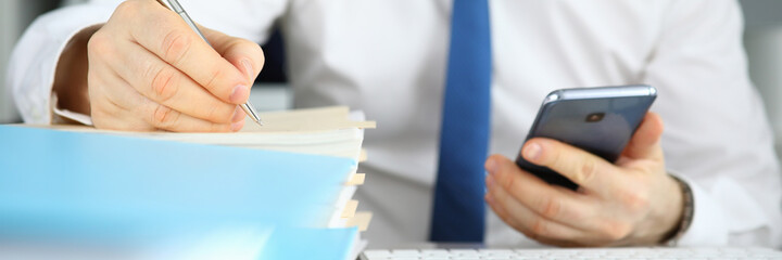 Close-up of businessman filling important document sitting on modern office. Man in presentable suit holding smartphone. Signing contract. Business and technology concept