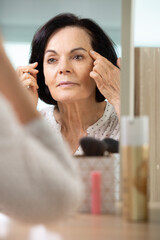 older woman pulls skin around her eye looking in mirror