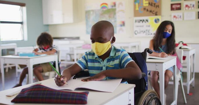Disable boy wearing face mask writing while sitting on his wheelchair in class