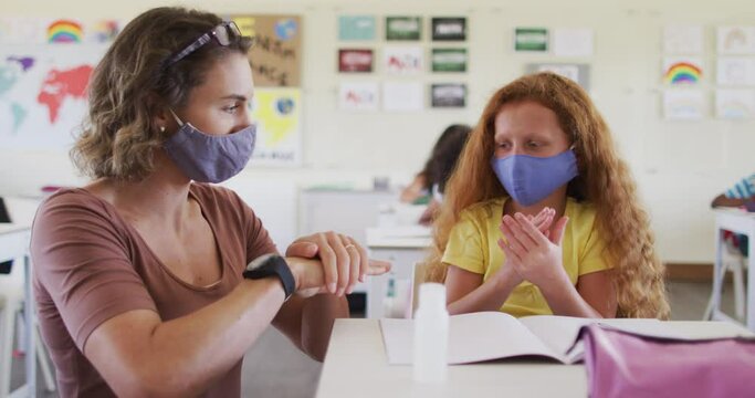 Female teacher and girl wearing face masks sanitizing her hands at school