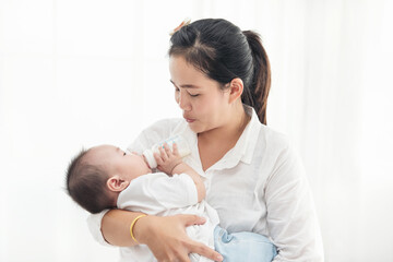 Obraz na płótnie Canvas Portrait of a happy asian mother giving bottle feeding to her baby in white bedroom. Baby is drinking milk from a bottle hold by the mother.