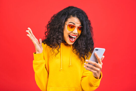Attractive curly american female with Afro hairstyle looks agitated at screen of smart phone, impressed by message content recieved from friend, has shocked surprised facial expression.