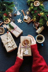 Top view of female hands in a red cozy sweater holding a decorated gift, Christmas decor, branches of a Christmas tree, and wrapped gifts on the table, celebrating and giving.