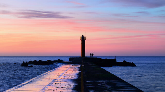 A couple of people standing near the orange lighthouse. Colorful sunset sky. Stunning cloudscape. Long exposure. National landmark, sightseeing, recreation theme. Baltic sea, Riga bay, Latvia