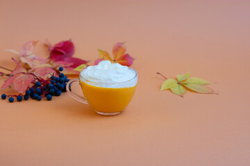 Pumpkin spice latte in a glass mug with whipped cream on  beige background. Autumn leaves. Hot autumn drink.