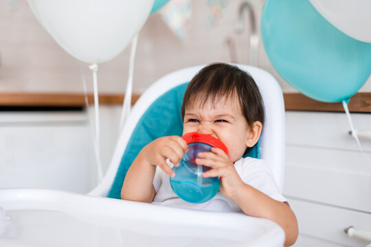 Little baby boy sitting in high chair at home on white kitchen and drinking water from sippy cup on background with balloons.