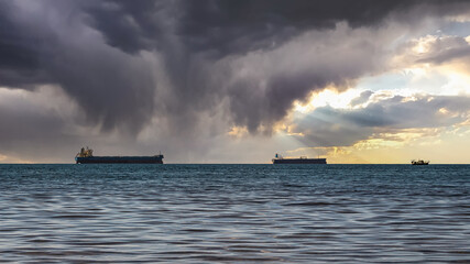 Close-up of a cloudy seascape and a bright sunset. Blue sea, storm clouds with sun rays over the horizon, and several cargo ships. Surroundings of the resort of Gelendzhik. Russia, Black sea coast