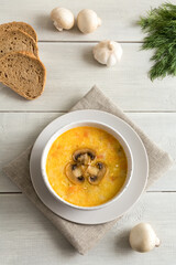 Mushroom soup in a white bowl on linen napkin. Whole champignons and bread flat lay on a wooden white background. Top of the soup is decorated with slices of sliced mushroom.