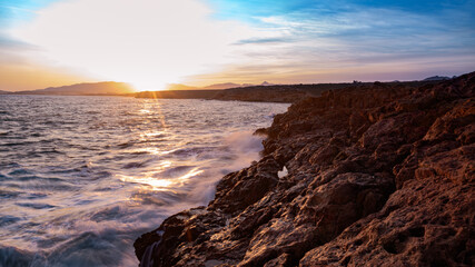 Fototapeta na wymiar Ocean landscape, rocks next to the sea and the waves crashing in the red rocks. Sunshine in the sunset with blue sky and the sun reflections on the sea.