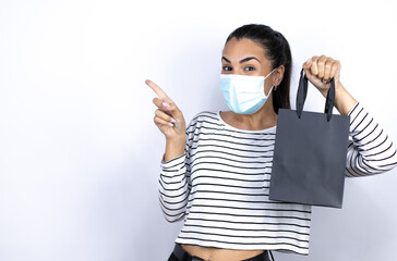 Young beautiful woman holding a black shopping bag wearing a medical mask to protect herself from the coronavirus pointing to the side . Black Friday