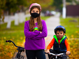 Brother and sister in the autumn park, riding bicycles wearing protective masks. In the background, a blurred autumn park. Pandemic and virus concept