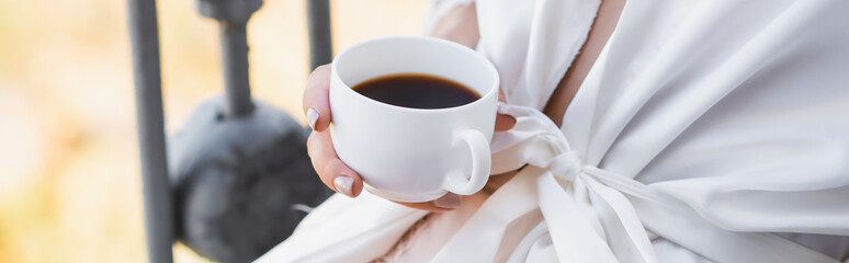 cropped view of woman in white robe drinking coffee on balcony, horizontal banner
