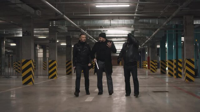 Full front-view shot of three assertive bandits in leather jackets leaving dark underground parking after crime taking off balaclava arguing and gesturing