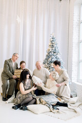 Happy multi generation family, sitting on the floor with pillows at stylish decorated light room, exchanging gifts in front of Christmas tree. Winter holidays, generations concept