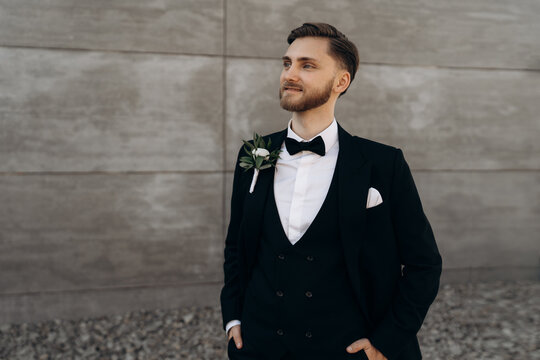 Handsome young groom with beard dressed in a classic black suit and white shirt and bow-tie sitting. Portrait of the groom on a background of a gray wall. Stylish man dressed in a black suit