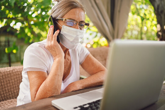 Senior woman face mask speak smartphone working online with laptop computer outdoor in garden