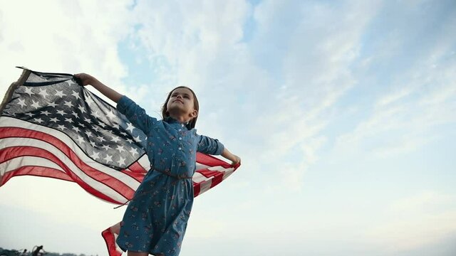 Patriotic female kid standing with American flag in hands against cloudy sky.