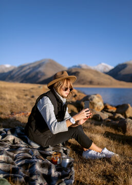 young beautiful girl on a picnic in nature, sitting on a plaid on the lake and taking pictures on the phone