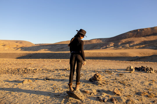 a young girl in black tight-fitting clothes with a beautiful figure stands on a stone in the middle of a huge desert with mountains at sunset