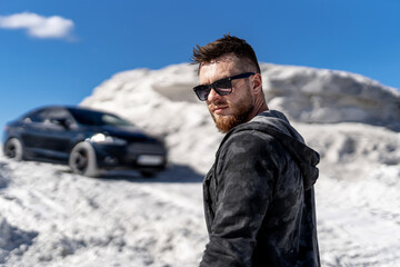 Portrait of a stylish bearded man in trendy sunglasses and leather jacket stands near black car. Fashion portrait on white rocks background.