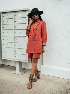 Woman in orange suit and black hat with braids standing by mailbox