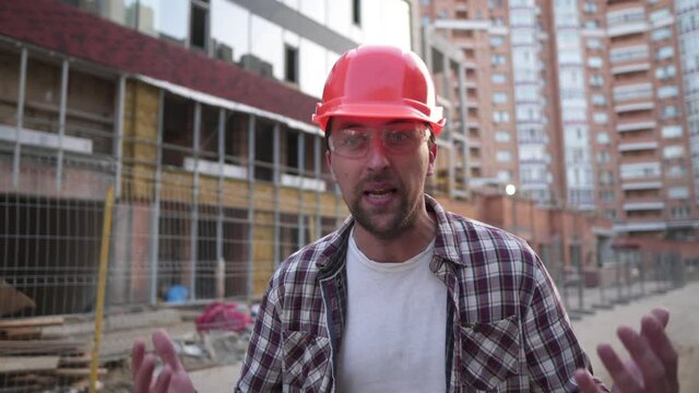 Young caucasian angry construction worker shouting at construction site. Angry engineer screaming. Evil foreman in hard hat shouts with his hands up. Hate, emotions and aggression concept