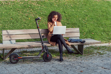 woman sitting on a bench talking on the phone with a laptop and an electric scooter