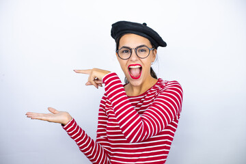 Young beautiful brunette woman wearing french beret and glasses over white background surprised, showing and pointing something that is on her hand