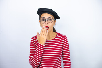Young beautiful brunette woman wearing french beret and glasses over white background surprised covering her mouth