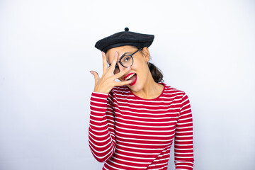 Young beautiful brunette woman wearing french beret and glasses over white background peeking in shock covering face and eyes with hand, looking through fingers with embarrassed expression