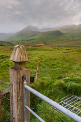 Old wooden gate pole in foreground and green field and mountains in the background. Low cloudy sky....