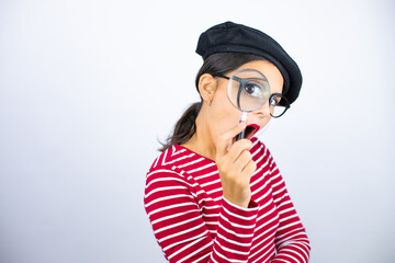 Young beautiful brunette woman wearing french beret and glasses over white background surprised looking through a magnifying glass