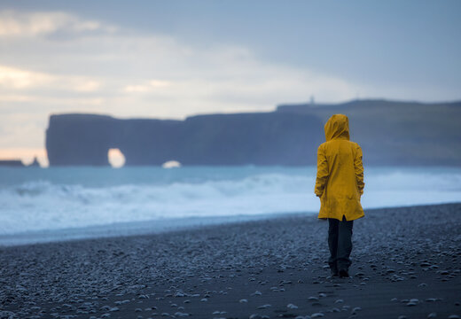 Girl in a yellow jacket walks on the black sand of a beach in iceland