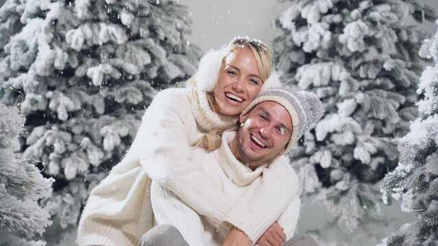 Happy young couple hugs and smiles and snow falls near christmas trees at the eve of new year celebration in winter day. Smiley man and woman weared white pullovers love each other