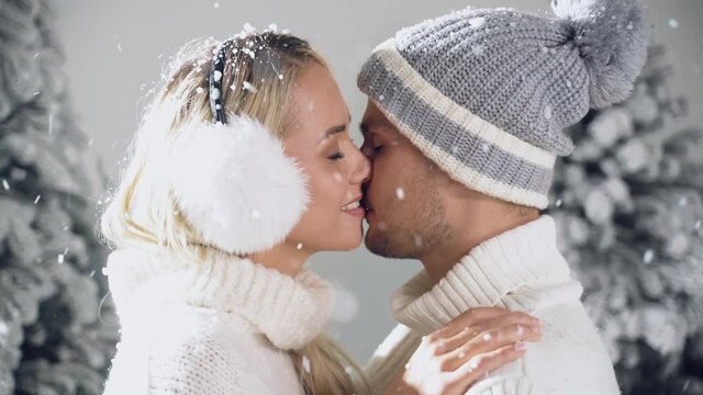 Falling snow and kisses. Happy young couple close-up hugs and kiss near christmas trees at the eve of new year celebration in winter day. Smiley man and woman weared white pullovers love each other