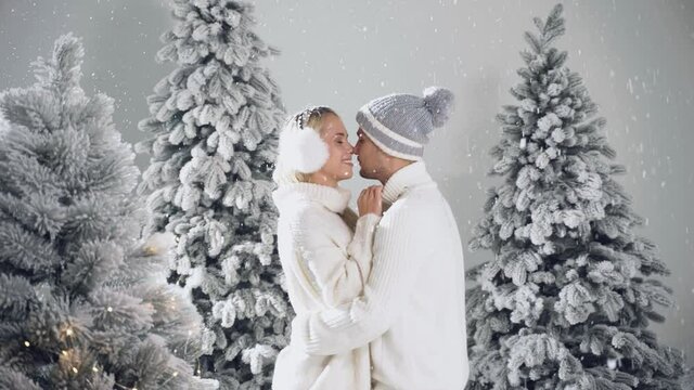 Falling snow and kisses. Happy young couple hugs and kiss near christmas trees at the eve of new year celebration in winter day. Smiley man and woman weared white pullovers love each other