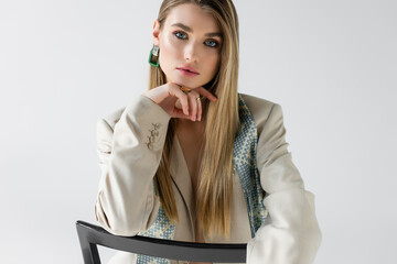 young woman in suit sitting on wooden chair and looking at camera on white