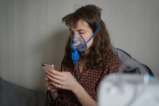 A young sick woman doing inhalation and using a phone at home