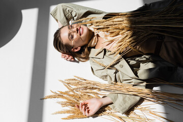 Obraz premium high angle view of cheerful woman in glasses, trench coat and scarf lying near wheat on white
